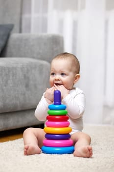 Cute baby girl playing with colorful toy pyramid sitting on carpet at home. Early development for kids.