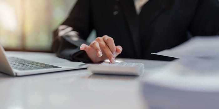 Business woman calculating monthly expenses, managing budget. Woman sitting at table using calculator to calculate tax refund, working at office with laptop on table.