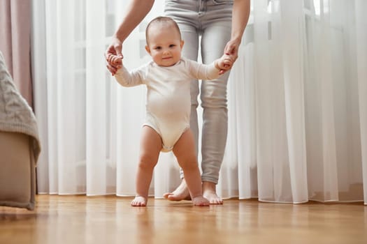 happy little baby girl learning to walk with mother help at home. child take her first steps