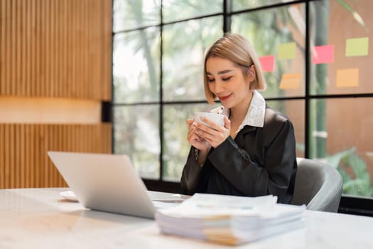 Smiling business woman drinking coffee while working on laptop from office.