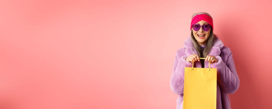 Fashionable asian senior woman going shopping in sunglasses and trendy winter clothes, holding paper bag from store and smiling happy at camera, pink background.