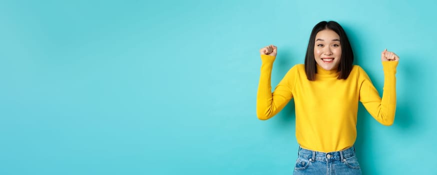 Hopeful asian girl winning prize, clench fists and smiling happy at camera, triumphing of achievement and success, standing over blue background.