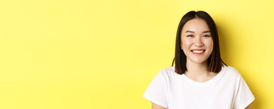 Beauty. Close up of smiling cute asian woman with perfect white smile teeth and glowing skin, standing over yellow background in t-shirt.