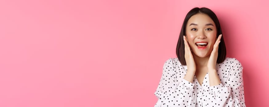 Close-up of beautiful asian woman touching clean skin face and staring at camera amazed, standing over pink background.