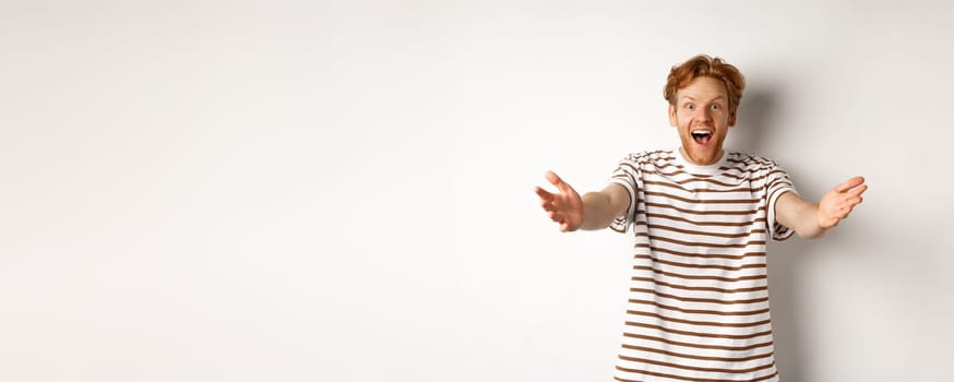 Cheerful man with red curly hair reaching hands forward, stretch out arms to welcome or congratulate you, smiling happy, standing over white background.