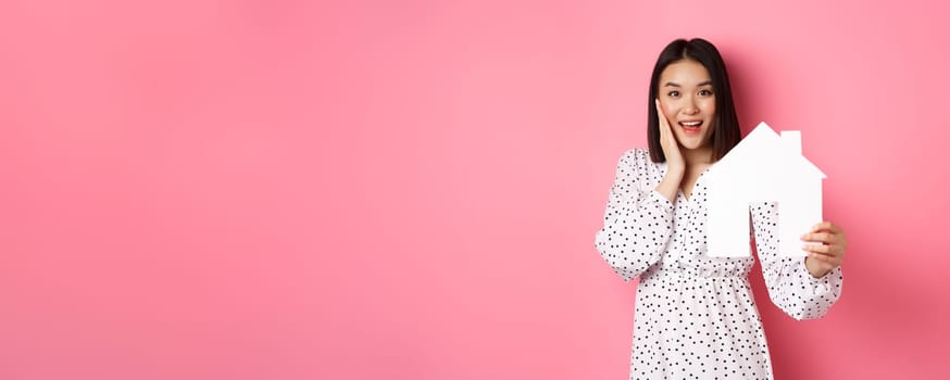 Real estate. Adult asian woman searching for home, holding house model and smiling, promo of broker company, standing over pink background.