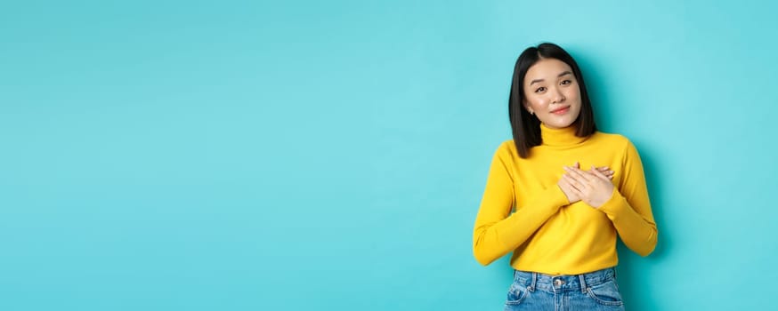 Portrait of beautiful heartfelt woman holding hands on heart, smiling and listening compassionate, standing over blue background in yellow pullover.