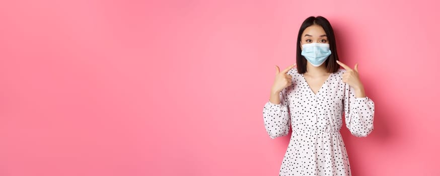 Coronavirus, social distancing and lifestyle concept. Cute asian woman pointing at face mask, asking to use measures against covid-19, standing over pink background.