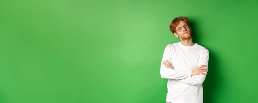 Confident young man with red hair, wearing glasses, looking smug at right side copy space, cross arms on chest self-assured, standing over green background.