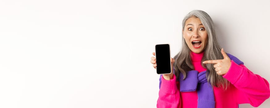 Online shopping. Beautiful asian grandmother smiling, pointing finger at smartphone blank screen, looking amazed, showing mobile application, standing over white background.