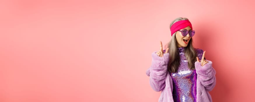 Hipster granny in sunglasses and glittering dress posing for photo with peace, victory signs, standing sassy against pink background.