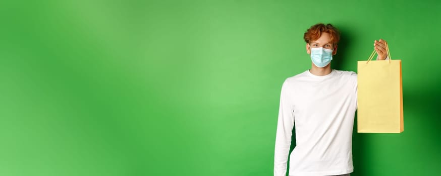 Portrait of handsome boyfriend in face mask giving shopping bag with valentines gift, smiling with eyes, standing over green background.