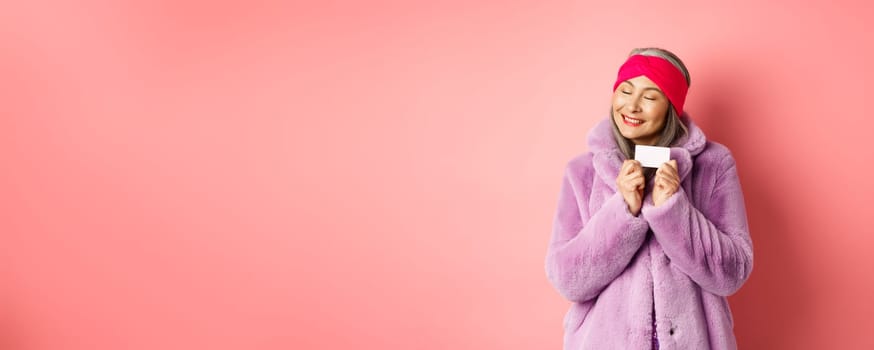 Shopping and fashion concept. Fashionable asian woman in purple faux fur coat, looking happy and showing plastic credit card, smiling cheerful, standing over pink background.