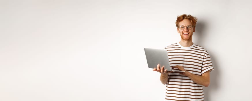 Smiling young man working on laptop and looking joyful, standing over white background.