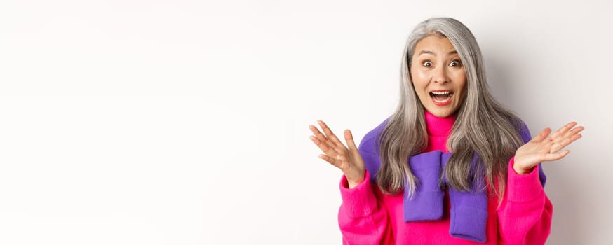 Close-up of excited asian senior woman spread hands sideways, gasping and looking amazed and surprised, hear great news, standing over white background.