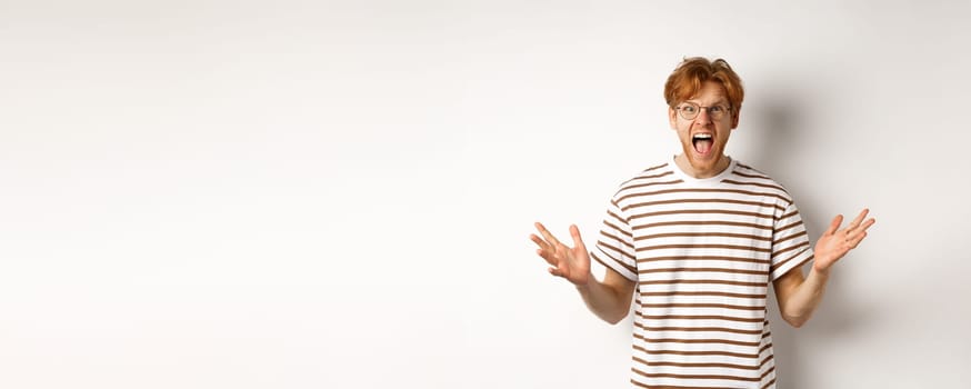 Frustrated man with red hair shaking hands and yelling at camera, looking angry and displeased, standing over white background.