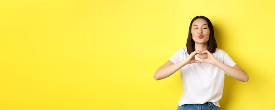 Beautiful asian woman showing I love you heart gesture, smiling at camera, standing against yellow background. Concept of valentines day and romance.