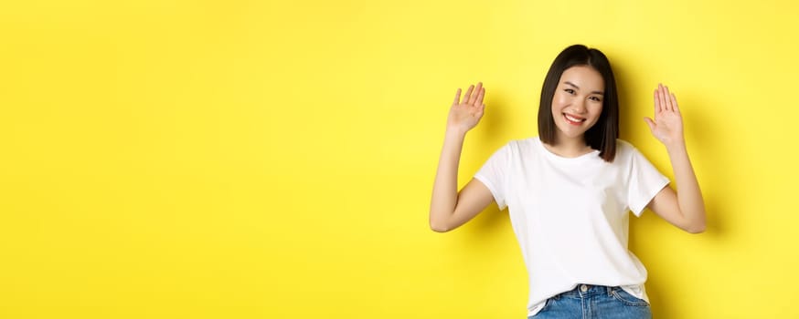 Friendly young asian woman saying hello, raising empty hands up and smiling, greeting you, standing over yellow background.