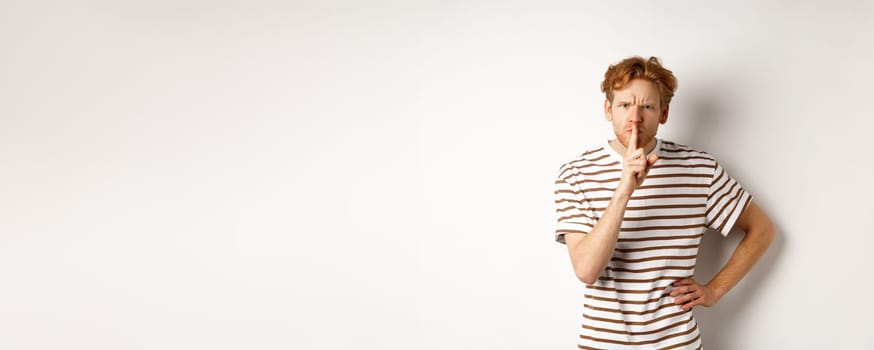 Angry redhead man looking displeased and hushing at camera, making shh gesture, standing over white background.