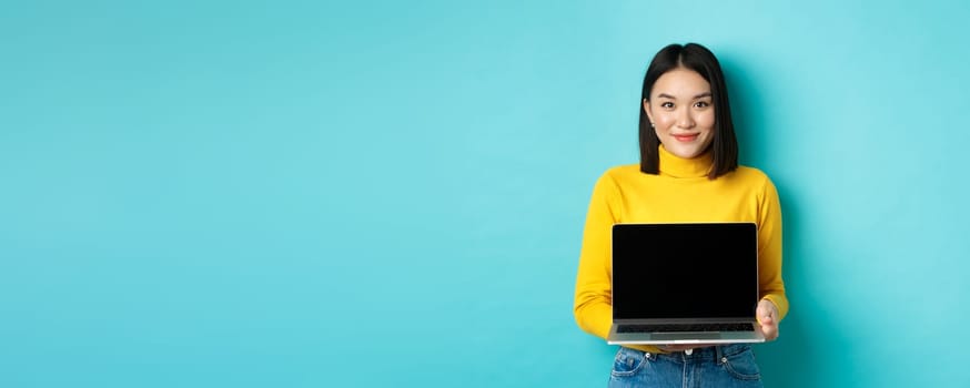 Beautiful and stylish asian woman demonstrate product on screen, showing empty laptop display and smiling, standing over blue background.
