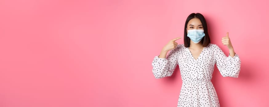 Coronavirus, social distancing and lifestyle concept. Cute asian woman pointing at face mask, showing thumb-up, support using preventive measures from covid-19, pink background.