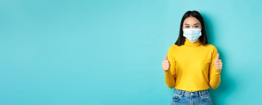 Covid-19, social distancing and pandemic concept. Supportive asian woman recommend promotion, wearing medical mask and showing thumbs up, standing over blue background.