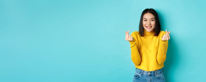 Beautiful asian girl in yellow sweater, smiling and showing finger hearts, standing happy against blue background.
