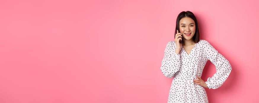 Beautiful asian woman having mobile conversation, making phone call and smiling, standing over pink background.