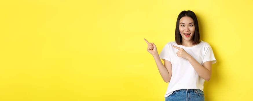 Beauty and fashion concept. Beautiful asian woman in white t-shirt pointing fingers left, standing over yellow background.