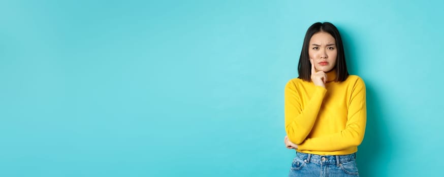 Image of pensive and serious asian woman touching chin, frowning and staring at camera puzzled, making difficult choice, standing over blue background.