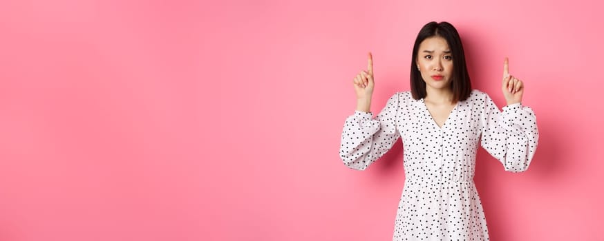Upset asian girl pointing fingers up, showing promo banner, staring at camera with disappointed gloomy look, standing in dress over pink background.