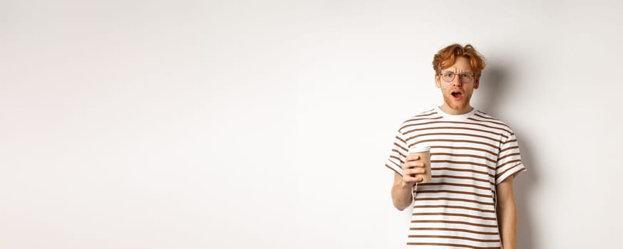 Amazed redhead man in glasses holding coffee cup and staring at camera with complete disbelief, standing in striped t-shirt against white background.