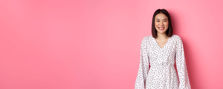 Happy korean woman in dress looking at camera, smiling and laughing with sincere expression, standing over pink background. Copy space