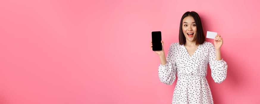 Cute asian woman shopping online, showing bank credit card and mobile screen, smiling and looking at camera, standing over pink background.