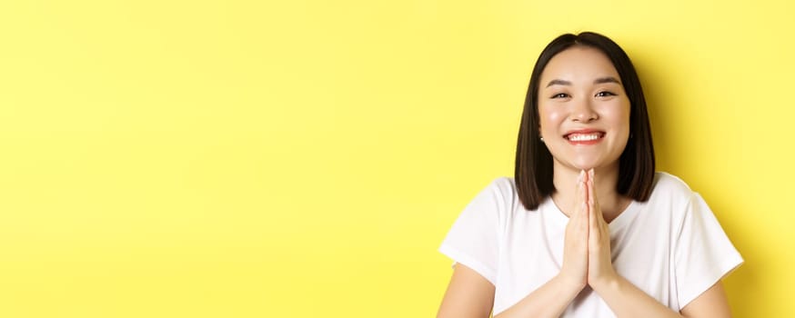 Close up of cute asian woman saying thank you and smiling, holding hands in namaste, pray gesture, standing over yellow background.