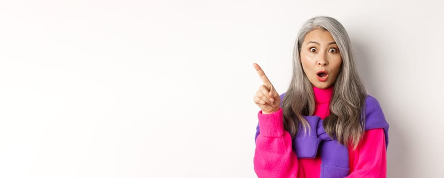 Fashion. Close up of amazed asian senior woman pointing finger upper left corner, gasping and looking impressed at camera, standing over white background.