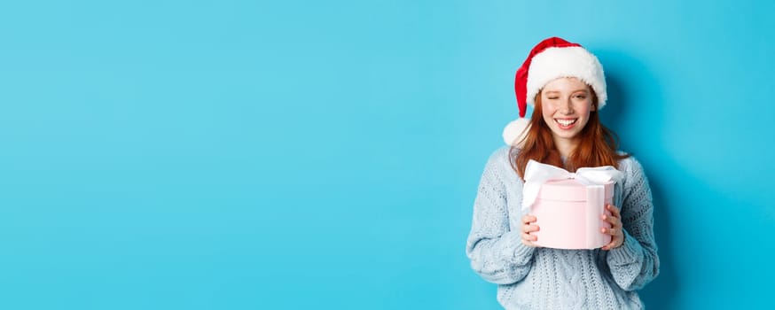 Winter holidays and Christmas Eve concept. Cute redhead girl in sweater and Santa hat, holding New Year gift and looking at camera, standing against blue background.