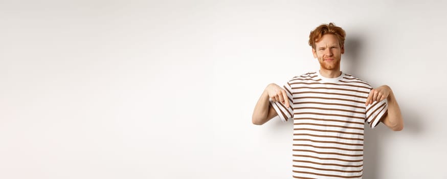 Disappointed redhead man shaking head displeased, showing something bad, pointing fingers down with skepical face, white background.