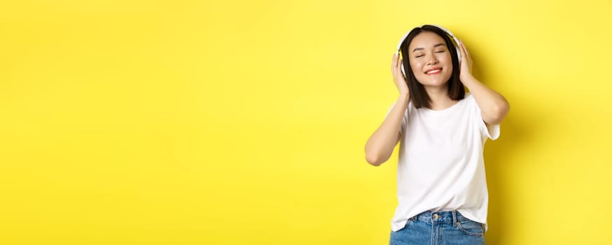 Attractive asian woman enjoying listening music in wireless headphones, smiling pleased and dancing with eyes closed, standing over yellow background.