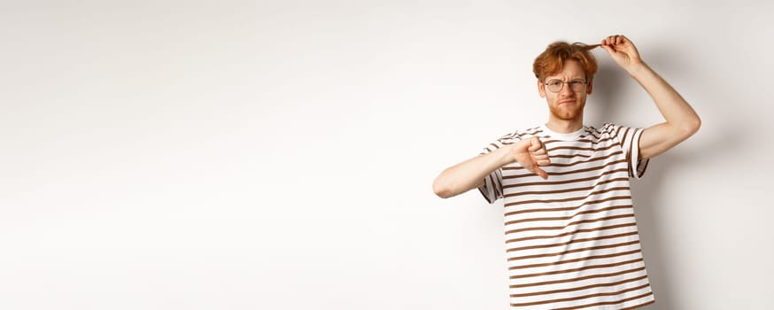 Young redhead man showing his messy haircut and thumbs-down, need hairdresser, standing over white background.