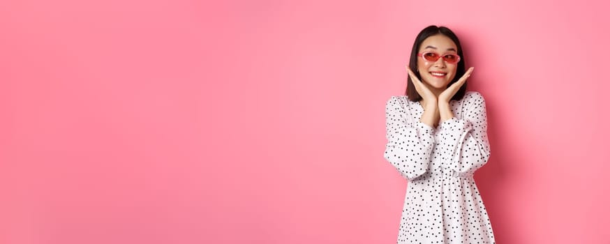 Tourism and lifestyle concept. Beautiful asian woman showing her clean cute face, wearing sunglasses, smiling and looking aside with admiration, standing over pink background.