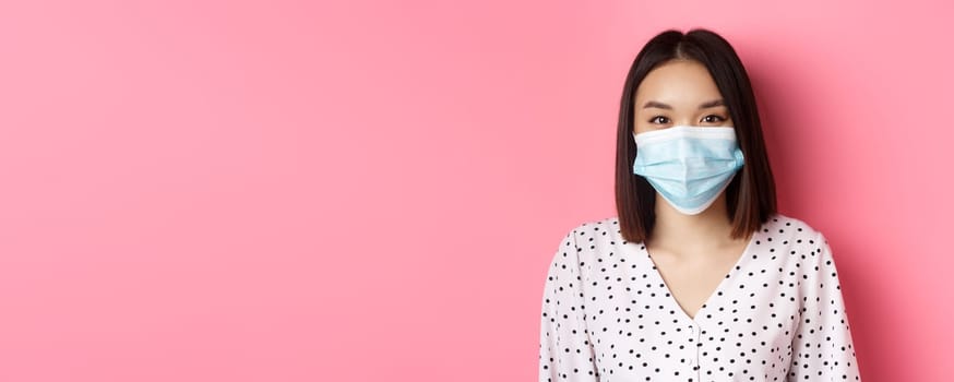 Covid-19, pandemic and lifestyle concept. Close-up of beautiful asian woman in face mask smiling with eyes, wearing protection from coronavirus in public, pink background.