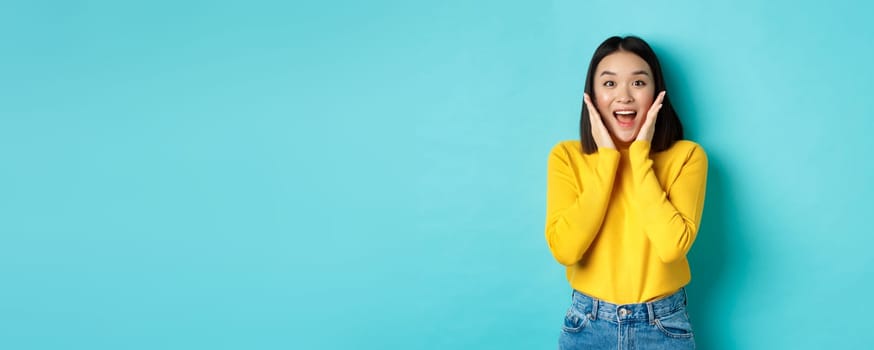 Portrait of pretty korean girl receive surprising news, looking amazed and happy at camera, standing over blue background.