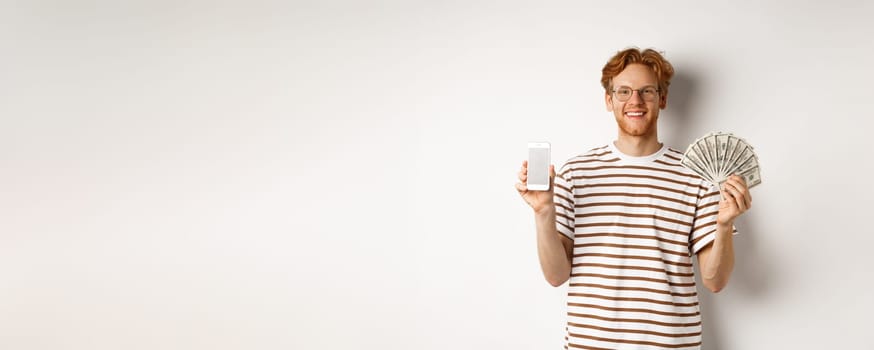 Smiling young redhead man in glasses showing smartphone blank screen and money, standing over white background.