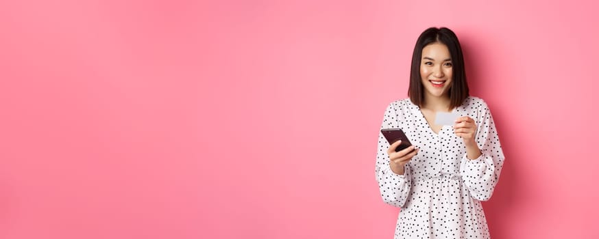 Beautiful asian woman enter credit card info in mobile phone app, shopping online, paying for order on smartphone, standing over pink background.