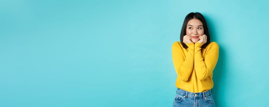 Beauty and fashion concept. Beautiful asian woman blushing and smiling, looking dreamy left, imaging something cute, standing against blue background.