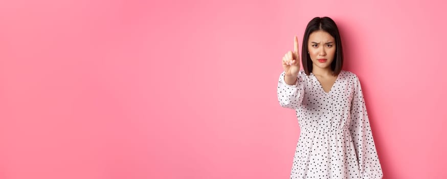Serious and angry asian woman telling to stop, frowning and showing finger in disapproval, prohibit something, standing in dress against pink background.