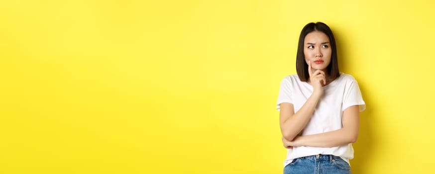 Beauty and fashion concept. Pensive asian woman thinking, looking thoughtful while pondering something, standing over yellow background.