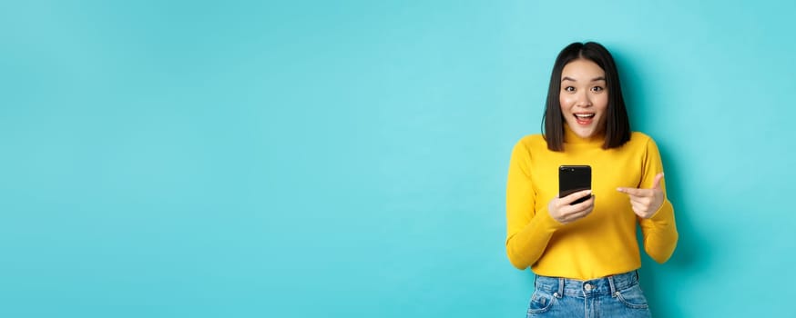 E-commerce and online shopping concept. Surprised asian woman demonstrate smartphone app, internet discounts, pointing finger at mobile phone, blue background.