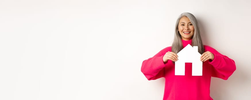 Real estate. Beautiful asian grandmother showing paper house and smiling happy, standing in pink sweater over white background.